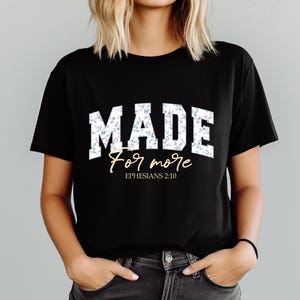 May include: Black t-shirt with the word "MADE" in white block letters filled with a blue floral pattern. Below, the words "For more" are written in a cursive font, with "Ephesians 2:10" below that.