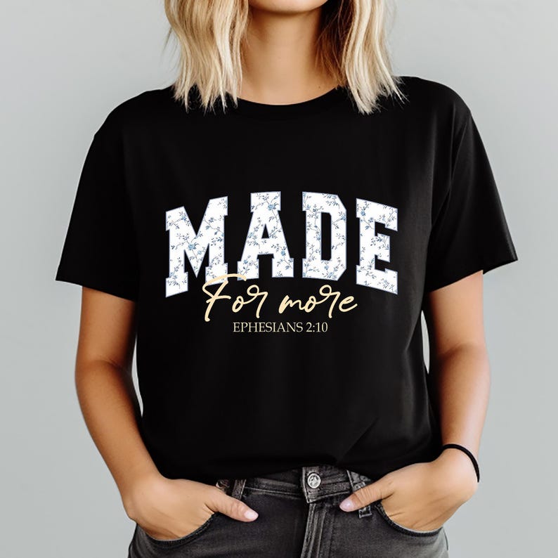 May include: Black t-shirt with the word "MADE" in white block letters filled with a blue floral pattern. Below, the words "For more" are written in a cursive font, with "Ephesians 2:10" below that.
