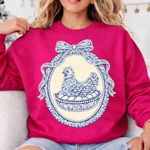 May include: A fuchsia sweatshirt features a blue and white floral hen design within an oval frame. The frame is adorned with a bow and pearl-like detailing. The hen is sitting on a nest of eggs.