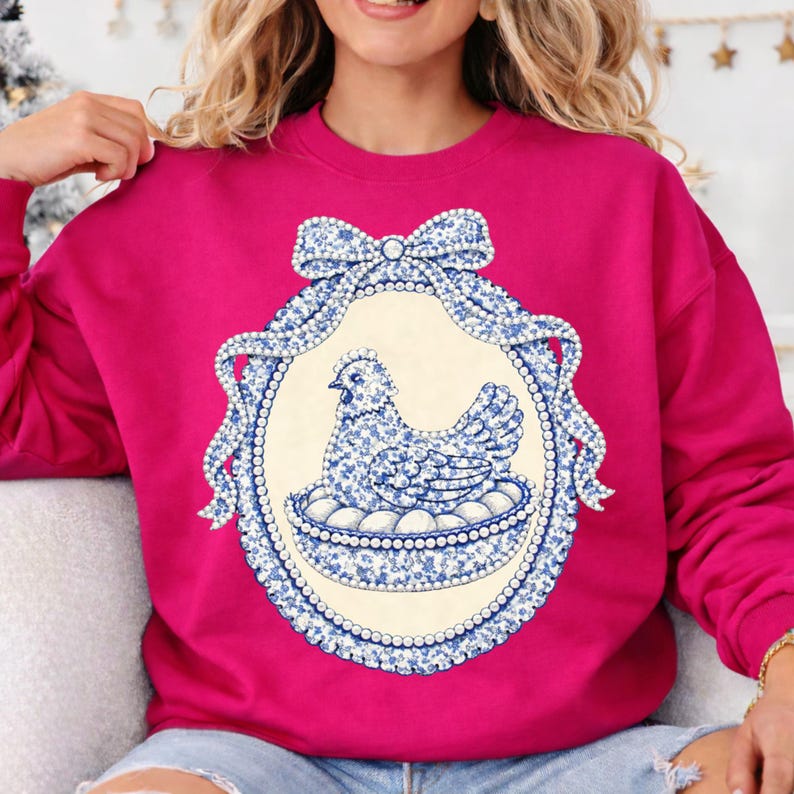 May include: A fuchsia sweatshirt features a blue and white floral hen design within an oval frame. The frame is adorned with a bow and pearl-like detailing. The hen is sitting on a nest of eggs.