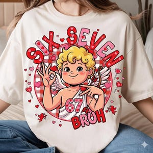 Puede incluir: Camiseta color crema con un diseño de Cupido de dibujos animados. El Cupido tiene cabello rubio rizado, alas y un arco y flechas. El diseño incluye el texto "SIX SEVEN BRUH" en rojo y rosa, rodeado de corazones.