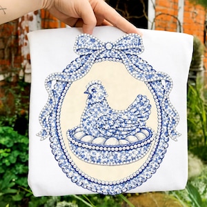 May include: White tote bag featuring a blue and white floral design of a hen sitting on eggs within an oval frame. The frame is adorned with a bow and pearl-like accents. The design is centered on the bag.