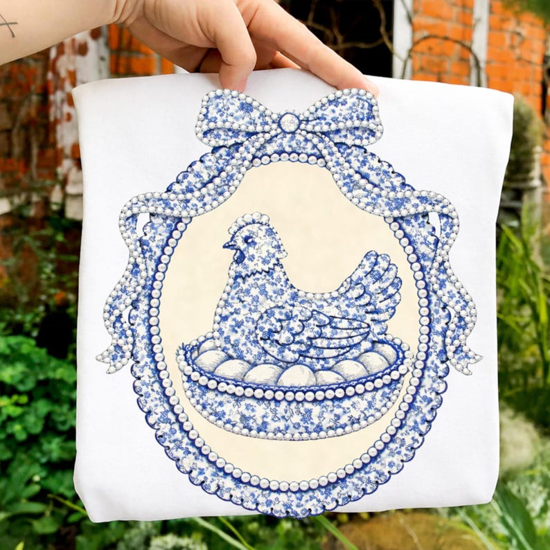 May include: White tote bag featuring a blue and white floral design of a hen sitting on eggs within an oval frame. The frame is adorned with a bow and pearl-like accents. The design is centered on the bag.