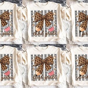 Puede incluir: Una colección de seis camisetas de color crema, cada una con un gráfico de un lazo con estampado de leopardo combinado con un lápiz. Los diseños incluyen el texto fourth grade, third grade, pre-k, kindergarten, first grade y second grade.