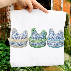 May include: A white tote bag featuring three decorative hen figurines. Each hen is perched in a basket adorned with faux pearls. The hens have blue floral patterns and are in different shades of blue and green.