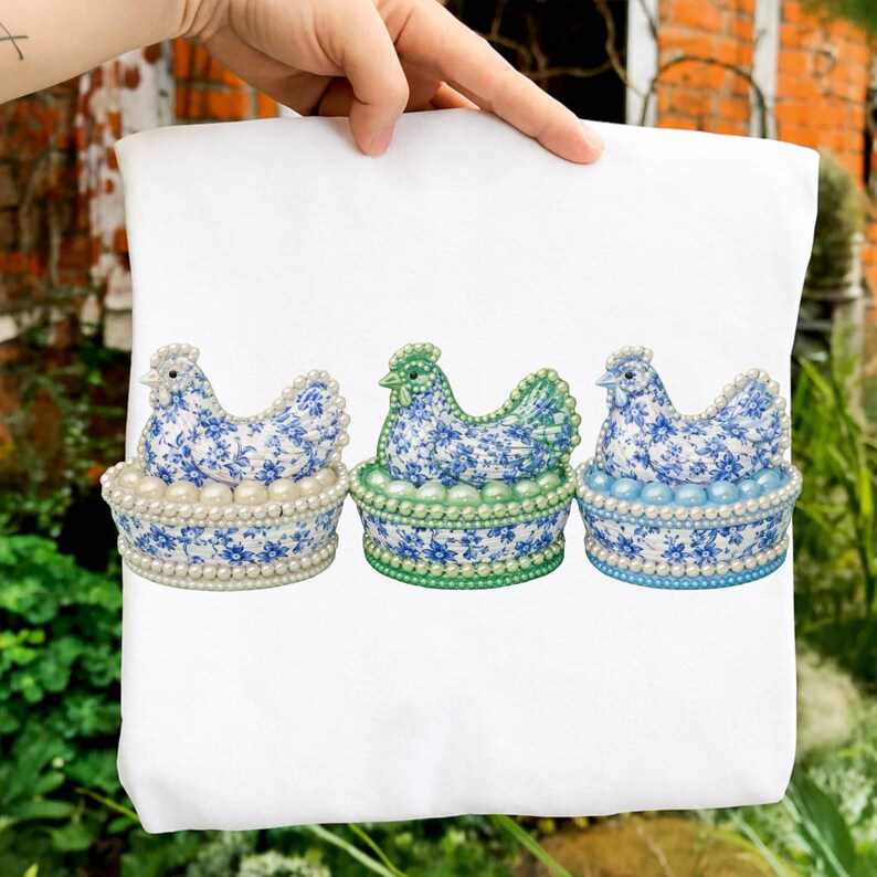 May include: A white tote bag featuring three decorative hen figurines. Each hen is perched in a basket adorned with faux pearls. The hens have blue floral patterns and are in different shades of blue and green.