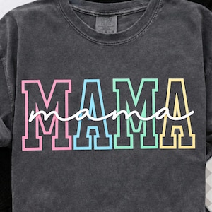 Puede incluir: Camiseta gris oscuro con la palabra "MAMA" en letras de bloque grandes y coloridas. La palabra "mama" está escrita en cursiva sobre las letras de bloque. La camiseta es de cuello redondo.