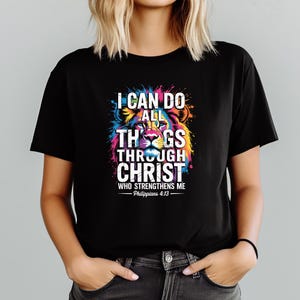 Może przedstawiać: Czarny t-shirt z kolorową grafiką lwa i napisem "I CAN DO ALL THINGS THROUGH CHRIST WHO STRENGTHENS ME - Philippians 4:13". Projekt lwa zawiera żywe odcienie niebieskiego, ż&oacute;łtego, r&oacute;żowego i zielonego.