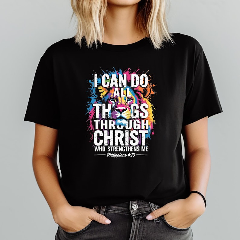 Może przedstawiać: Czarny t-shirt z kolorową grafiką lwa i napisem "I CAN DO ALL THINGS THROUGH CHRIST WHO STRENGTHENS ME - Philippians 4:13". Projekt lwa zawiera żywe odcienie niebieskiego, ż&oacute;łtego, r&oacute;żowego i zielonego.
