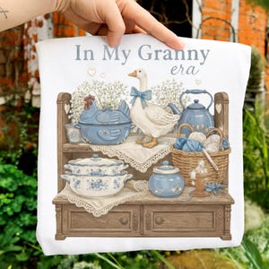 Puede incluir: Paño de cocina blanco con el texto "In My Granny era". El diseño presenta un ganso con un lazo azul, utensilios de cocina azules y una cesta de hilo en una estantería de madera. El paño tiene una estética vintage y rústica.