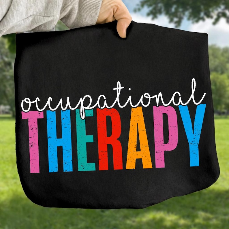 May include: Black tote bag with the words "occupational THERAPY" in white script and colorful block letters. The bag is held up against a green background.