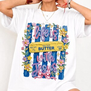 Puede incluir: Camiseta blanca con un diseño gráfico colorido. El diseño incluye las palabras "life is with Jesus" en rosa y azul, una barra de mantequilla amarilla y detalles florales. La camiseta tiene cuello redondo y mangas cortas.