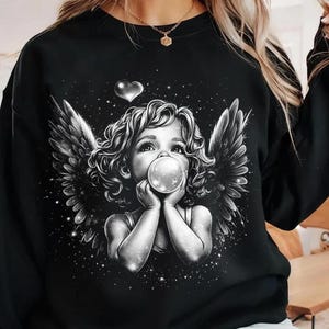 Puede incluir: Sudadera negra con una ilustración en escala de grises de una figura angelical soplando una burbuja. El diseño incluye alas de ángel, un corazón y un fondo estrellado. La obra de arte es detallada y caprichosa.