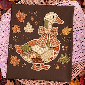 Puede incluir: Un panel de tela marrón oscuro con una aplicación central de ganso en patchwork decorada con un gran lazo a cuadros. El ganso está rodeado de hojas de otoño, bellotas y ramitas bordadas, todo el diseño está delineado con pequeñas cuentas blancas.