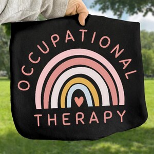 May include: Black tote bag featuring a colorful rainbow design with the words "OCCUPATIONAL THERAPY" in pink. The rainbow includes layers of pink, white, yellow, and blue, with a small pink heart at the bottom. The bag is held up against a green background.