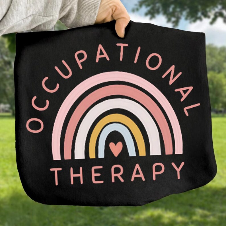 May include: Black tote bag featuring a colorful rainbow design with the words "OCCUPATIONAL THERAPY" in pink. The rainbow includes layers of pink, white, yellow, and blue, with a small pink heart at the bottom. The bag is held up against a green background.