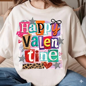 Puede incluir: Camiseta color crema con un gráfico colorido de "Happy Valentine". El diseño presenta letras recortadas en varios colores, estrellas, corazones y un lazo. La camiseta tiene un corte holgado.