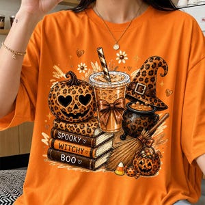 Puede incluir: Una camiseta naranja con un estampado gráfico de una calabaza con estampado de leopardo, un vaso con pajita, un sombrero de bruja, un caldero, una escoba y tres libros apilados con el texto SPOOKY, WITCHY y BOO en los lomos de los libros.