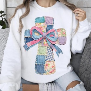 Puede incluir: Sudadera blanca con un diseño de cruz de retazos y un lazo grande. La cruz está hecha de varios patrones de tela, incluyendo floral, mezclilla y cuadros. El lazo es rosa y azul, agregando un toque de color al diseño.