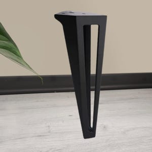 May include: A single black metal furniture leg with a triangular design. The leg has a flat top with pre-drilled holes for mounting and a tapered shape with a rectangular cutout. The leg is designed to provide support and stability.