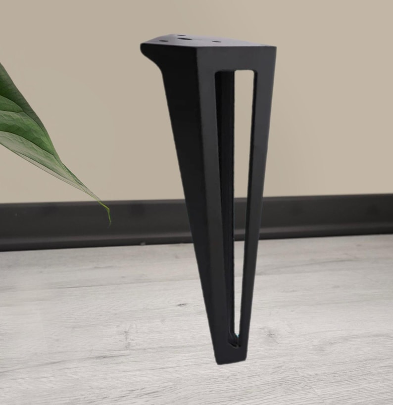 May include: A single black metal furniture leg with a triangular design. The leg has a flat top with pre-drilled holes for mounting and a tapered shape with a rectangular cutout. The leg is designed to provide support and stability.