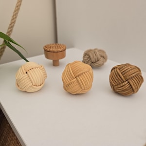 Może przedstawiać: Kolekcja dekoracyjnych węzłów i małego drewnianego gałki. Węzły wykonane są z tkanych włókien naturalnych w odcieniach beżu i brązu. Gałka ma okrągłą górę i krótką, cylindryczną podstawę. Przedmioty ułożone są na białej powierzchni.