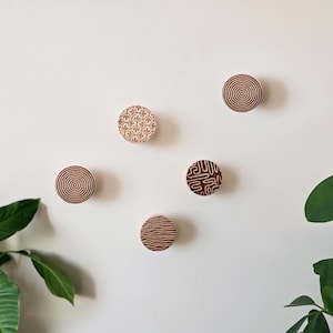May include: Five round wooden wall hooks with different geometric patterns. The hooks are brown and have a natural wood finish.