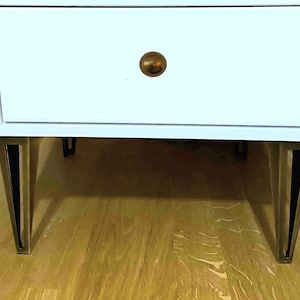 May include: White nightstand with two drawers, featuring a gold-colored knob and angled legs with gold-colored accents. The nightstand is set on a wooden floor.