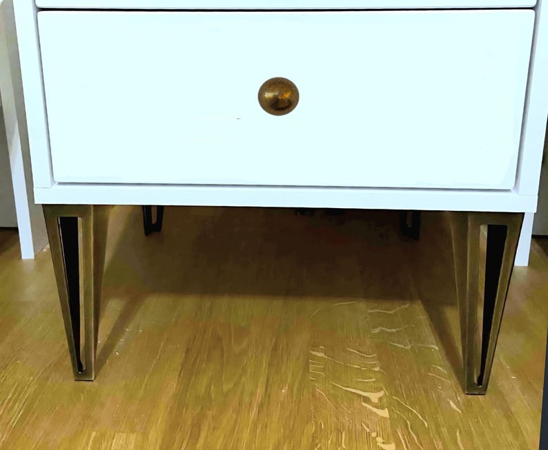 May include: White nightstand with two drawers, featuring a gold-colored knob and angled legs with gold-colored accents. The nightstand is set on a wooden floor.