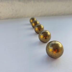 Może przedstawiać: Sześć metalicznych, złotych kul o teksturowanej powierzchni, ułożonych po przekątnej na białej powierzchni. Kule są ostre, z rozmytym tłem. Kule wydają się być dekoracyjne, być może do dekoracji wnętrz.