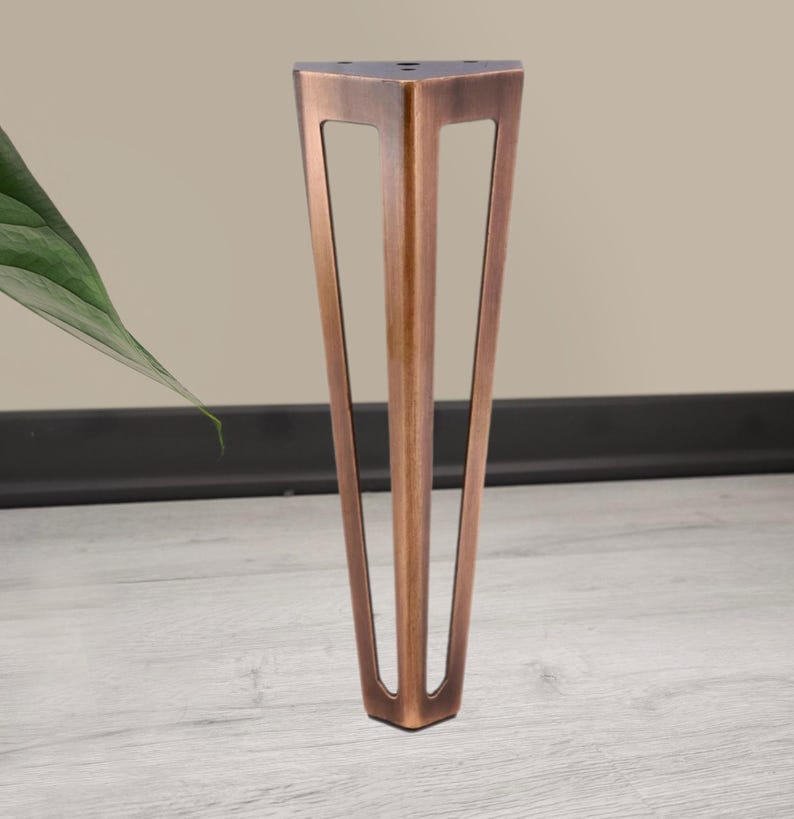 May include: A bronze-colored furniture leg with a triangular top and three angled, open-sided supports. The leg is made of metal and has a matte finish. The design is modern and geometric, suitable for various furniture styles.