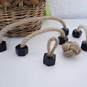 Puede incluir: Una colección de adornos decorativos de cuerda. Los adornos incluyen lazos de cuerda, nudos y piezas de cuerda arqueadas unidas a bases hexagonales negras. Una cesta tejida se encuentra en el fondo.
