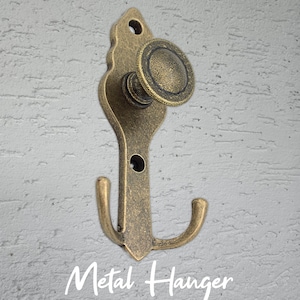 Może przedstawiać: Antyczny metalowy wieszak z mosiądzu z ozdobną g&oacute;rą i zakrzywionym haczykiem. Wieszak ma okrągłą gałkę i napis "Metal Hanger" w ozdobnej czcionce.