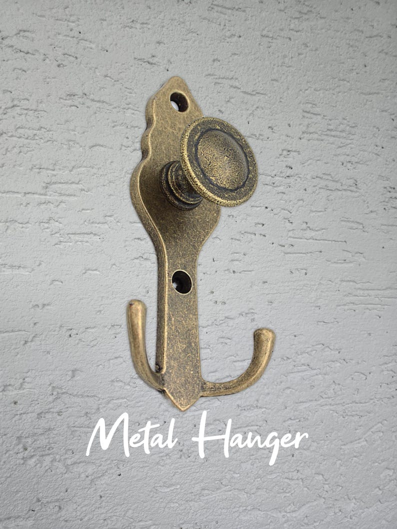 Może przedstawiać: Antyczny metalowy wieszak z mosiądzu z ozdobną g&oacute;rą i zakrzywionym haczykiem. Wieszak ma okrągłą gałkę i napis "Metal Hanger" w ozdobnej czcionce.
