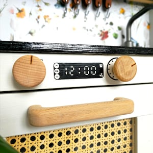 May include: White toy oven with a black top and a wooden handle. The oven has a digital display showing 12:00 and two wooden knobs.