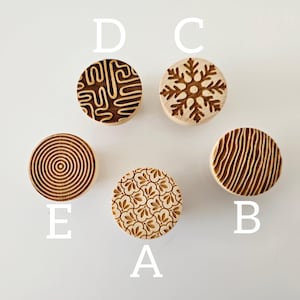 Puede incluir: Cinco sellos de madera con diferentes diseños. Los sellos están etiquetados de la A a la E. Los diseños incluyen un copo de nieve, un patrón de espiral, un patrón de hojas, un patrón de líneas onduladas y un patrón geométrico.