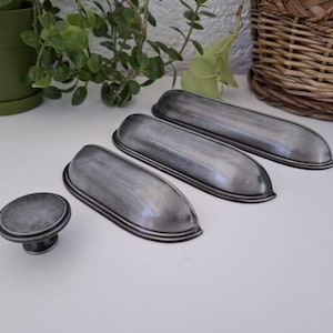 May include: A set of four antique-style cabinet hardware pieces. The set includes three elongated pulls and one round knob, all with a distressed silver finish and black trim. The hardware is arranged on a white surface.
