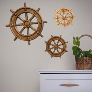 Puede incluir: Tres decoraciones de pared de timón de madera de diferentes tamaños están montadas en una pared gris claro. Una pequeña cesta tejida con asa contiene una planta verde. Las ruedas son marrones y doradas.