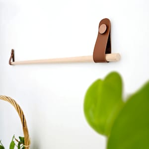 Puede incluir: Un toallero de madera con correas de cuero marrón y un botón de cobre. La barra está montada en una pared blanca.