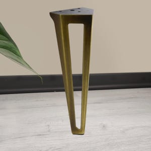 May include: A single, angled furniture leg in a brass finish. The leg has a triangular top with pre-drilled holes and a rectangular cutout in the center. The leg is positioned on a light wood-grain surface.