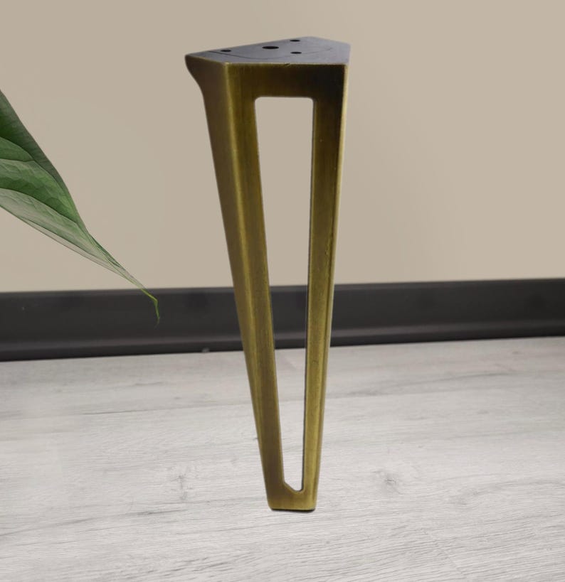 May include: A single, angled furniture leg in a brass finish. The leg has a triangular top with pre-drilled holes and a rectangular cutout in the center. The leg is positioned on a light wood-grain surface.