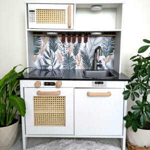 May include: White play kitchen with a black countertop, a black stovetop with a digital timer that reads "12:00", a white oven with a woven rattan door, a white sink with a chrome faucet, and a white cabinet with a wooden handle. The backsplash is a tropical leaf patterned wallpaper.