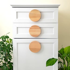 May include: White chest of drawers with three drawers and wooden round knobs. The chest of drawers has a simple design and is painted white. The knobs are made of natural wood and add a touch of warmth to the piece.