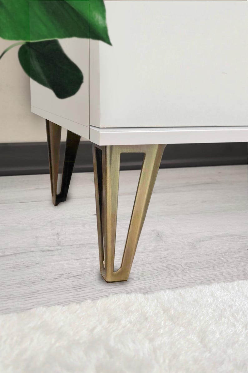May include: A white cabinet with brass-colored, angled legs. The legs are a distinctive feature, contrasting with the white body of the cabinet. The design suggests a modern or mid-century aesthetic. A green plant leaf is visible on the left side.