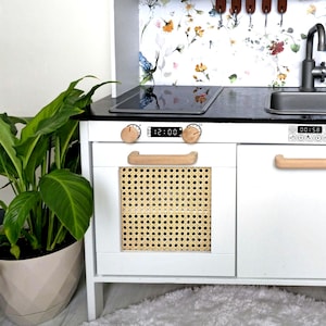 May include: White toy kitchen playset with a black stovetop, a white oven with a woven rattan door, and a white cabinet with a wooden handle. The kitchen has a floral wallpaper backdrop.