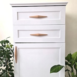 Può includere: Comò bianco con tre cassetti e una porta dell'armadio. I cassetti e la porta dell'armadio hanno maniglie di legno a forma di diamante.