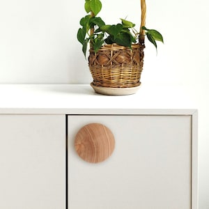 Może przedstawiać: Okrągły, jasnobrązowy gałka do szafki wykonana z drewna. Gałka ma lekko fakturowaną powierzchnię. Gałka jest zamontowana na białych drzwiach szafki.