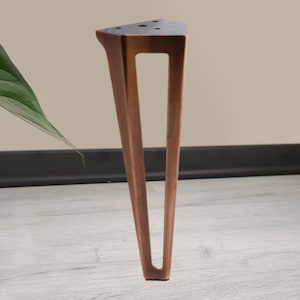 May include: A single, angled wooden furniture leg with a triangular top and a rectangular cutout. The leg is a warm brown color and stands on a light gray wooden floor. The background is a neutral beige.