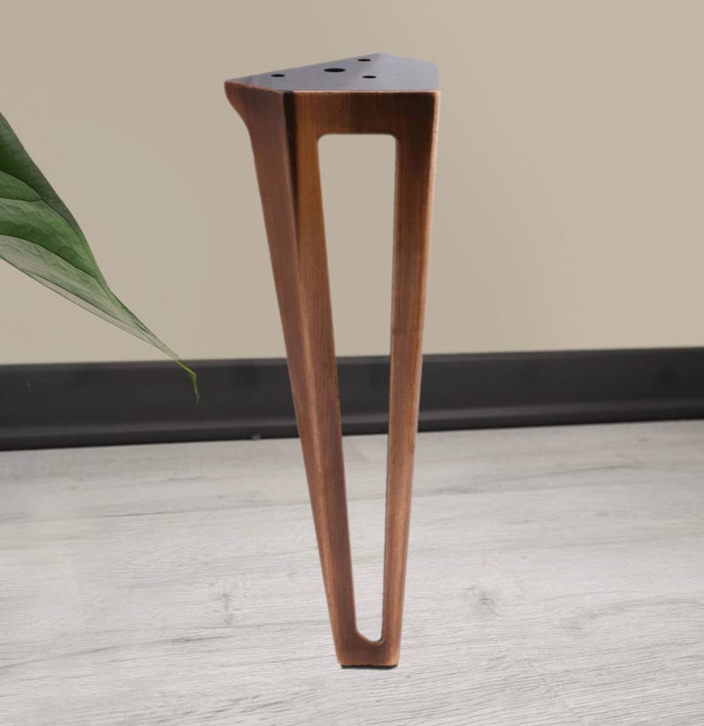 May include: A single, angled wooden furniture leg with a triangular top and a rectangular cutout. The leg is a warm brown color and stands on a light gray wooden floor. The background is a neutral beige.