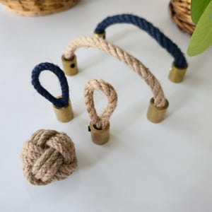 May include: A collection of nautical-themed hardware, including rope handles and loop pulls. The handles are made of natural rope with brass-colored metal ends. One handle is navy blue, and the other is beige. A decorative rope knot is also included.